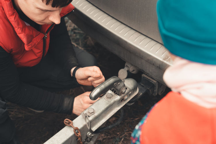 Man connects a trailer to the towbar of his car, and a child watches the process. 