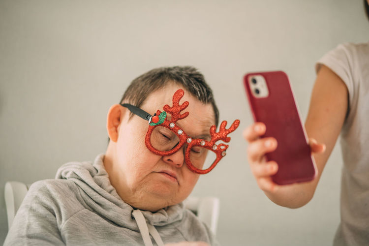 Girl helps sleepy elderly woman with down syndrome in funny christmas glasses,  video call by phone