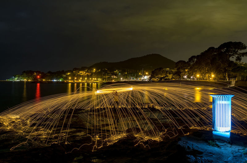 Illuminated light trails by river against sky at night