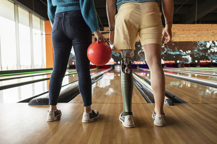 Woman holding ball with friend wearing artificial limb standing on hardwood floor at bowling alley