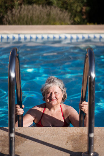 Positive senior female in swimwear going down in swimming pool and holding stainless handrails while relaxing in sunny day