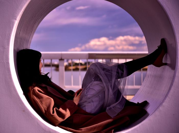 Side view of woman relaxing on circle shape structure against sky during sunset
