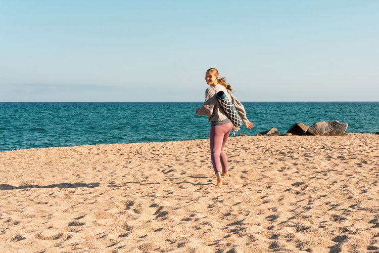 Full body side view of young happy barefoot female in activewear with rolled yoga mat looking at distance over the shoulder to the camera while standing on sandy beach near sea