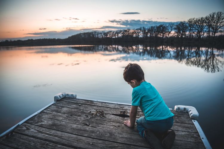 Boy on jetty over lake against sky during sunset