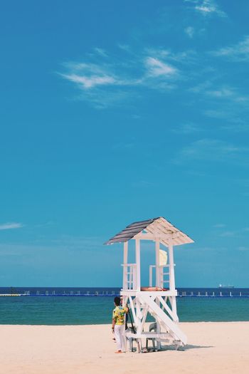 Rear view of man standing by lifeguard hut at beach against sky