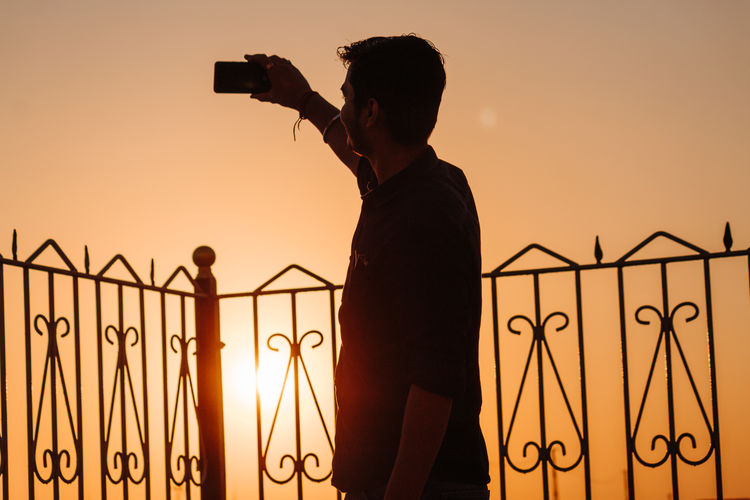 Man taking selfie on smart phone by railing against sky during sunset