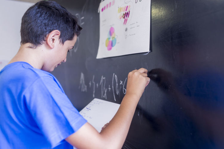Side view of schoolboy writing on blackboard in classroom