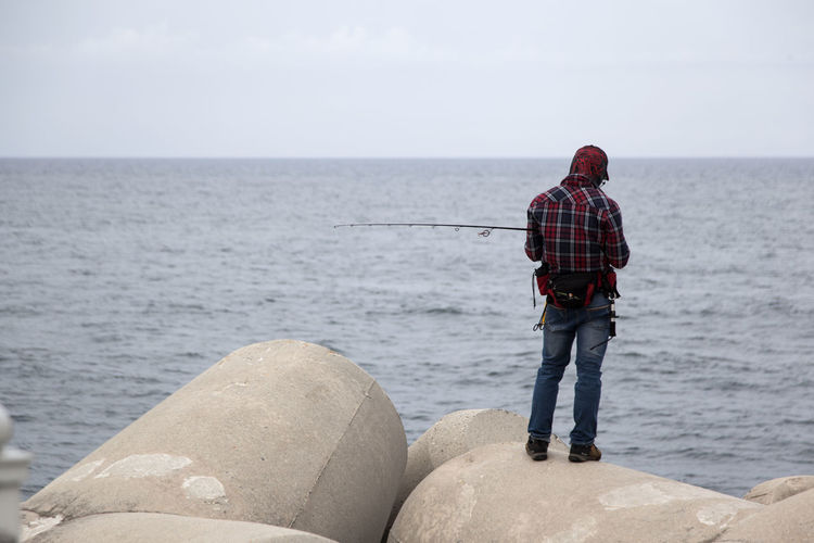 Rear view of man standing on rock while fishing at sea against sky