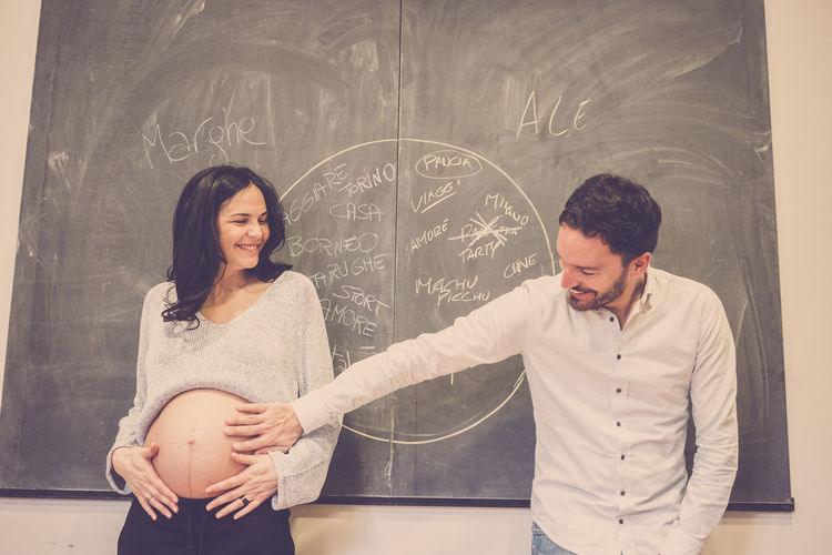 Smiling man touching belly of pregnant wife against blackboard