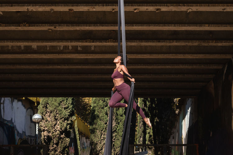 Slender african american female balancing on hanging hammock while doing aerial yoga above ground under bridge in city
