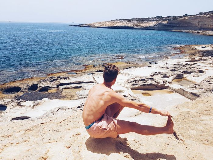 High angle view of shirtless man practicing pistol squat on rock looking at sea