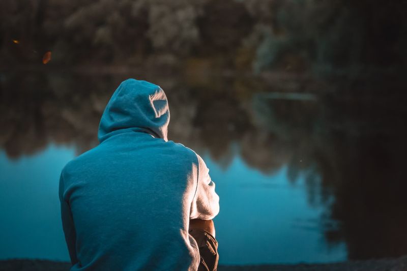 Rear view of man wearing hooded shirt sitting by lake during dusk