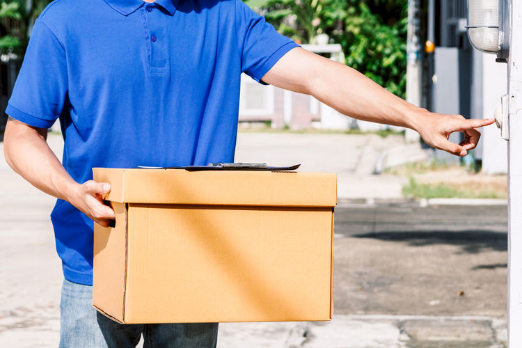 Midsection of delivery person holding cardboard box while ringing doorbell outdoors