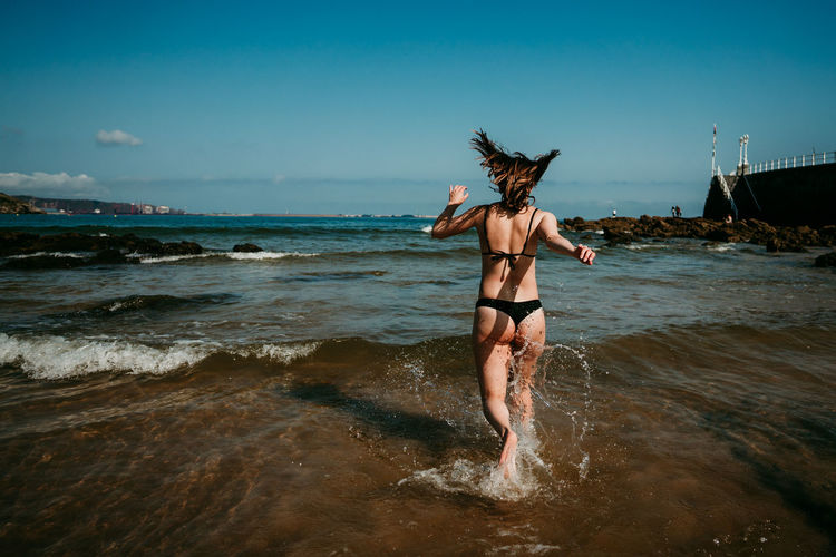 Back view of delighted female in bikini running in shallow water of sea while having fun and enjoying summer vacation