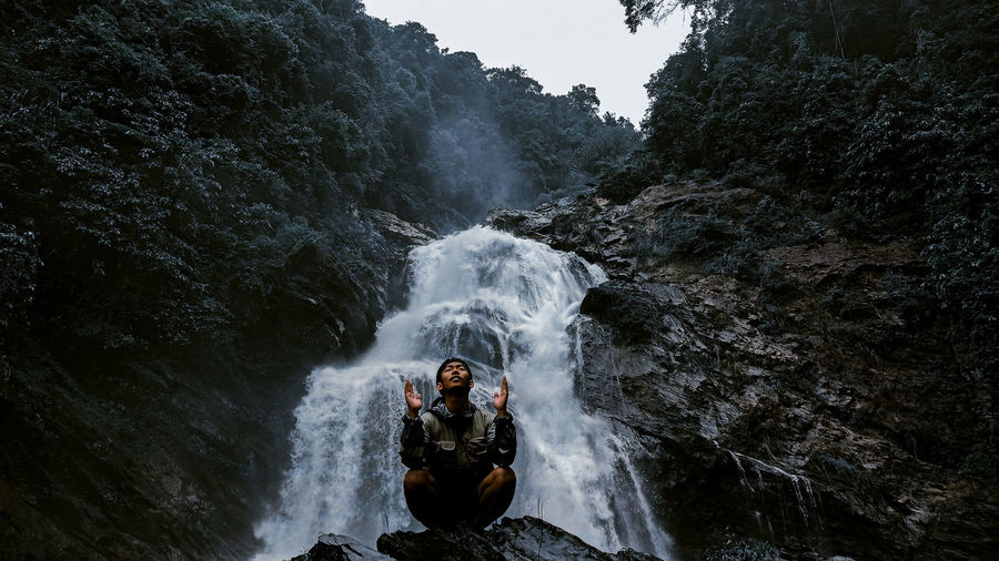 Young man meditating while sitting cross legged on rock against waterfall at forest