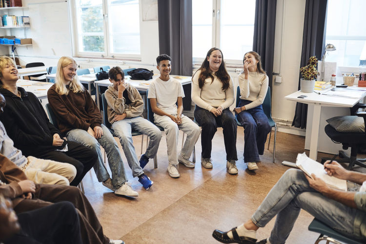 Happy male and female students sitting with counselor in group therapy at school