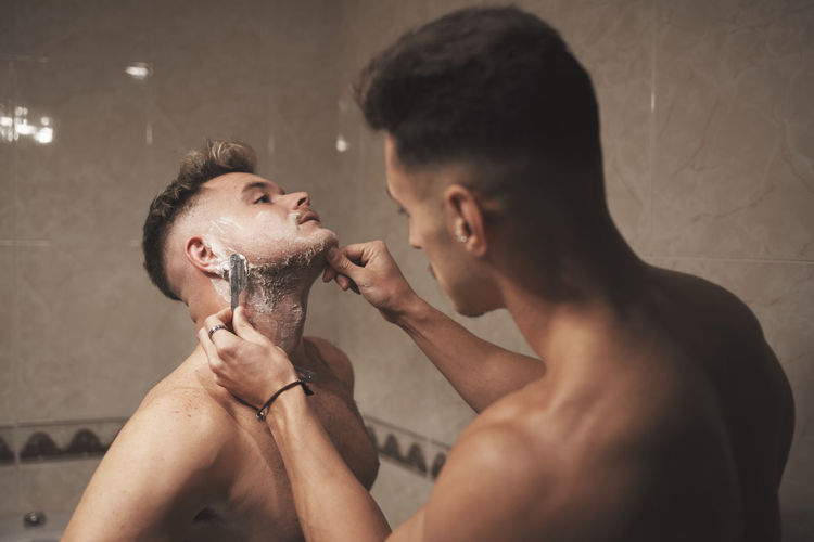 A young man is shaving his boyfriend with a razor in the bathroom