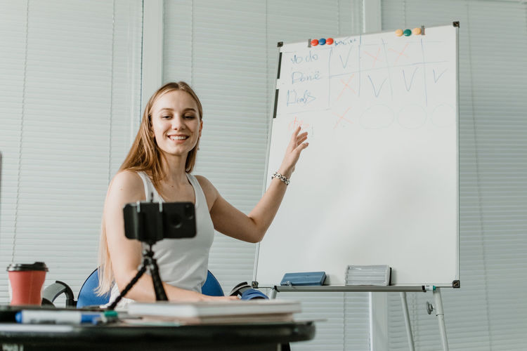 Smiling young woman explaining over whiteboard while blogging with smart phone at home