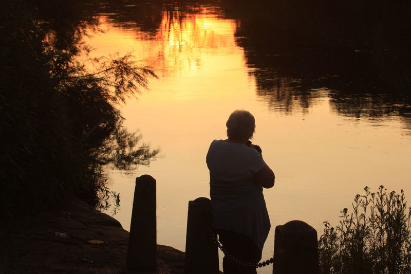 Rear view of woman sitting by lake against sky during sunset