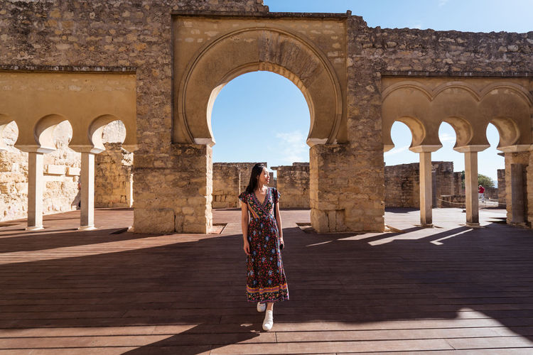 Full body of asian female traveler standing near stone ancient portico with columns and arched passages during trip in cordoba town