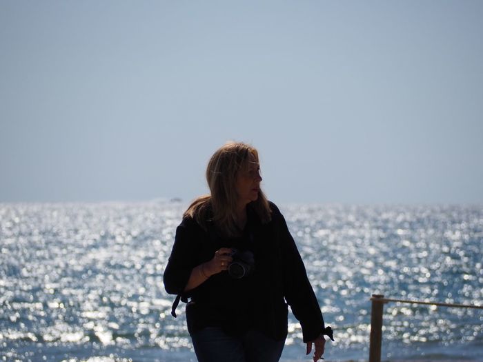 Mature woman holding camera while standing against sea at beach