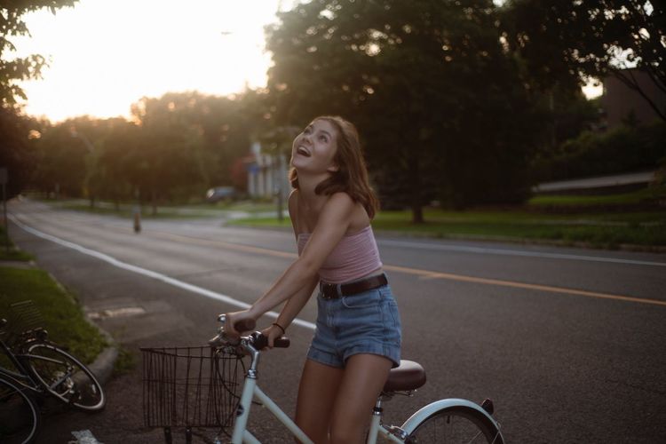 Cheerful young woman riding bicycle on road during sunset