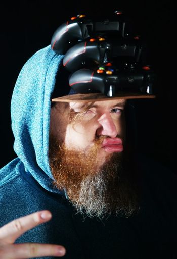 Portrait of bearded man with stacked game controllers on cap against black background