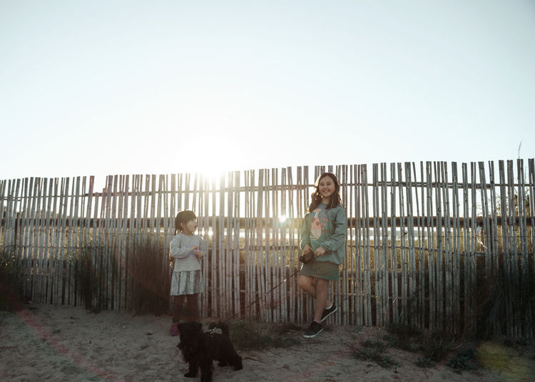 Rear view of two girl standing by fence with dog against clear sky