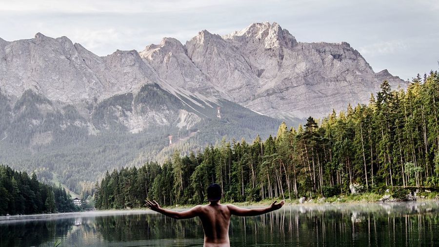 Rear view of shirtless man standing by lake against mountains