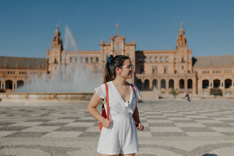 Young tourist woman wearing a white dress is looking away and smiling while standing in seville.