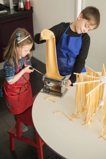 From above of little girl with older brother using pasta machine while preparing homemade noodles in home kitchen