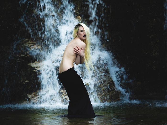 Full length of shirtless woman standing against waterfall