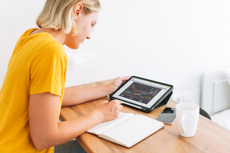 Young woman in yellow t-shirt studies cryptocurrency charts and stock quotes on digital tablet