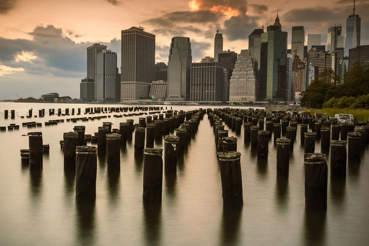 Wooden posts in river against buildings in city during sunset