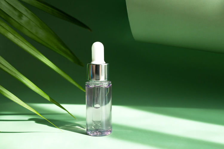 A transparent cosmetic bottle with a pipette next to a palm leaf in the rays of the bright sun