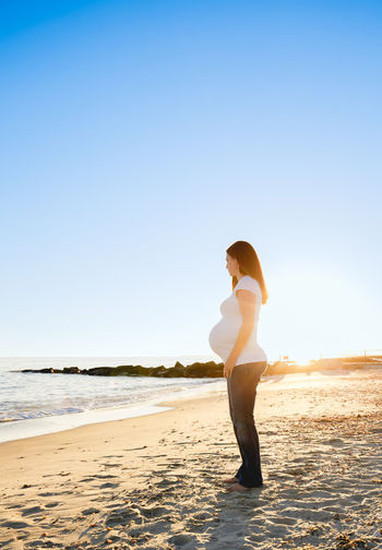 Full length of young pregnant woman 3rd trimester standing at beach against sunset clear sky