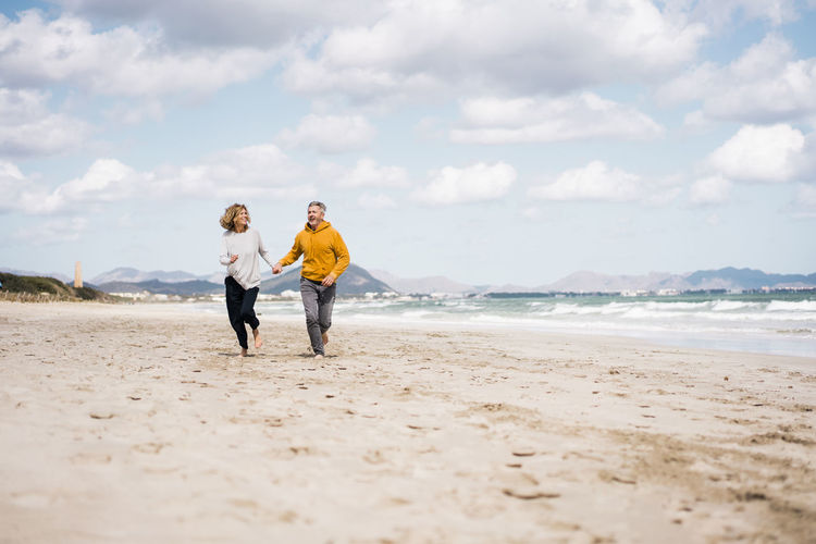 Cheerful mature couple holding hands running on shore at beach