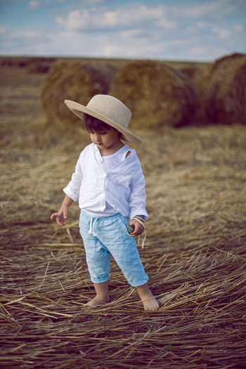 Boy a child in a straw hat and blue pants stands in a mowed field with stacks in the summer