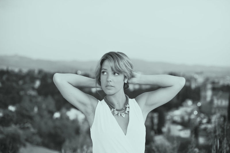 Beautiful young woman with hands behind head against city