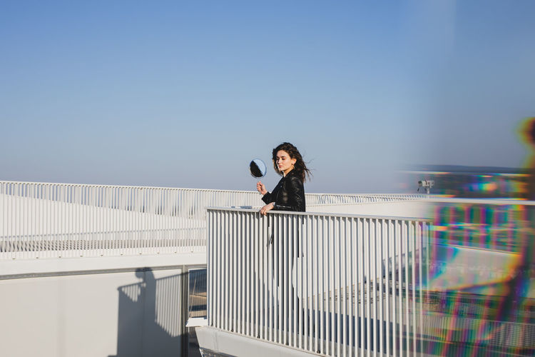 Side view of young woman holding mirror looking away while standing on terrace against clear blue sky