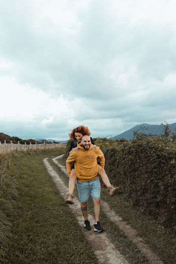 Happy hipster man giving playful girlfriend piggyback ride while walking on rural road between green farm and sea coast in cloudy day