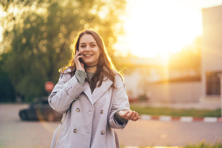 Smiling beautiful girl with long hair in a grey trench coat using smartphone in the spring 