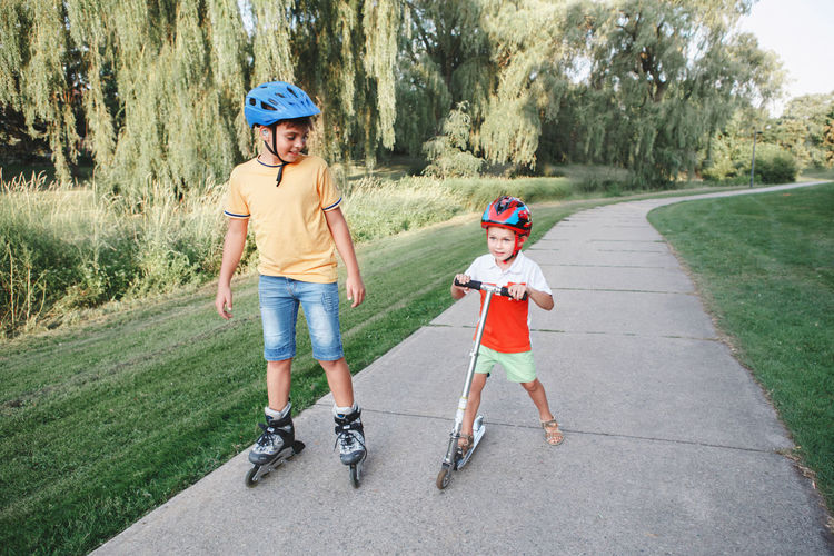 Caucasian boys brothers in helmets riding roller skates and scooter. siblings friends relationships.