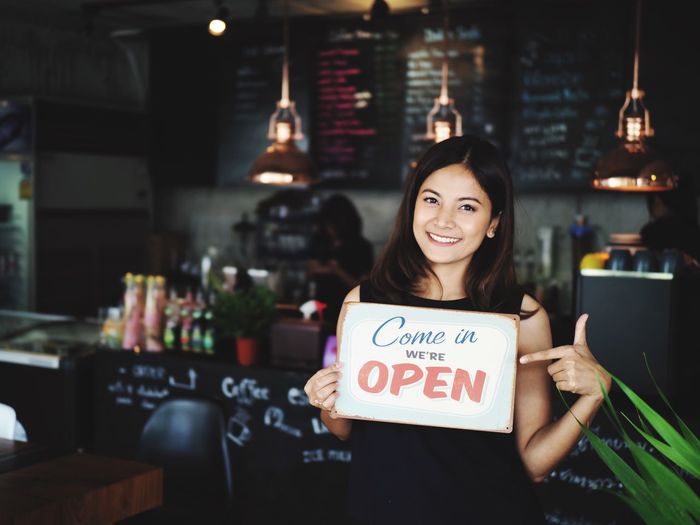 Portrait of smiling young woman holding open sign while standing in cafe
