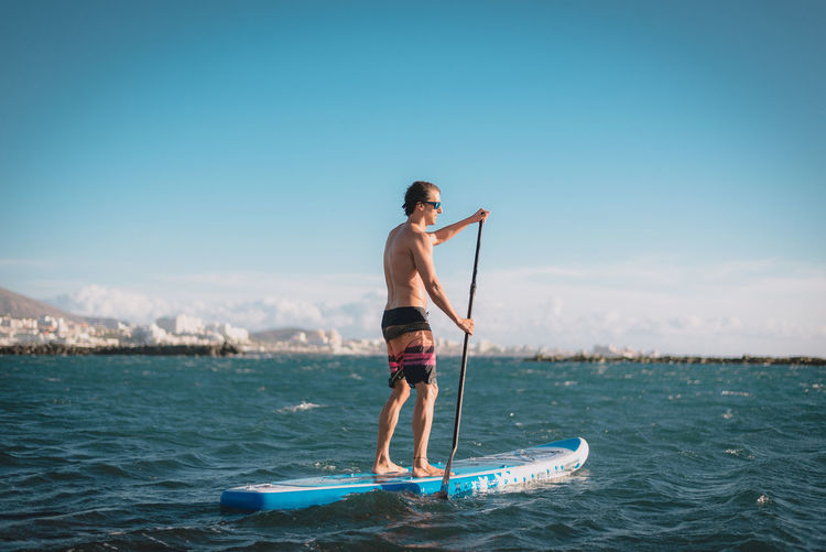 A young sportsman man practices paddle surfing on the beach under a blue sky