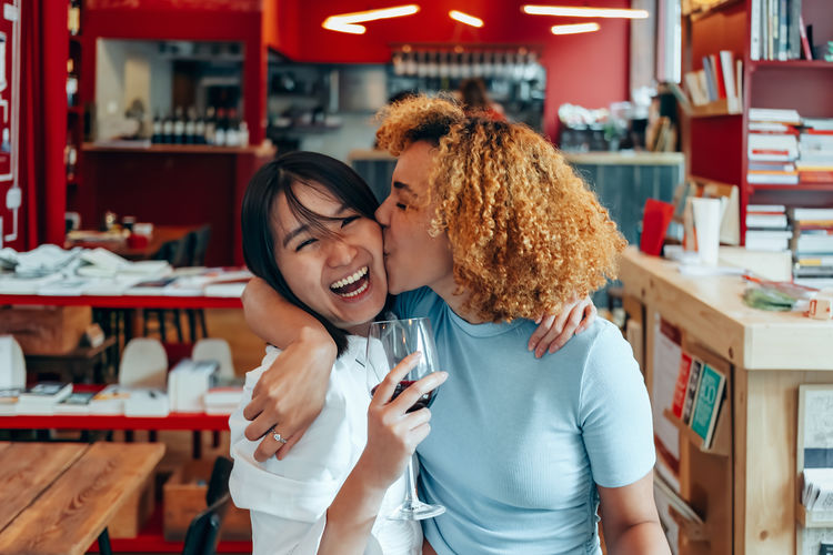 Girlfriends at the cafe hug each other and laugh - brazilian girl kisses asian woman on the cheek