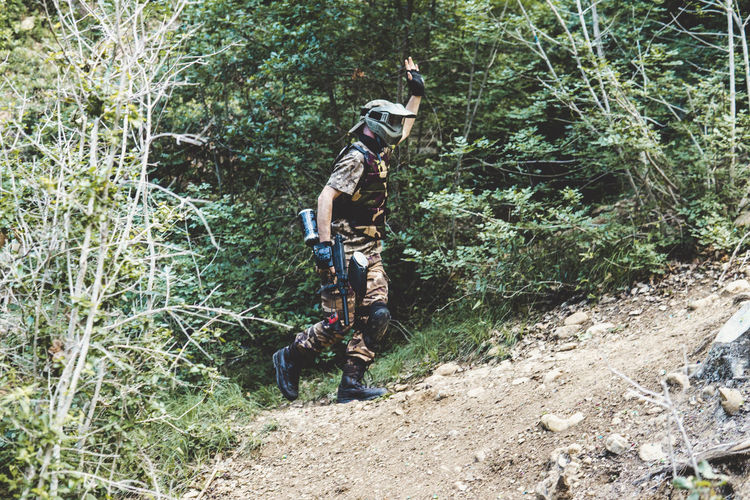 Side view of army solider with gun waving hand while walking on hill in forest
