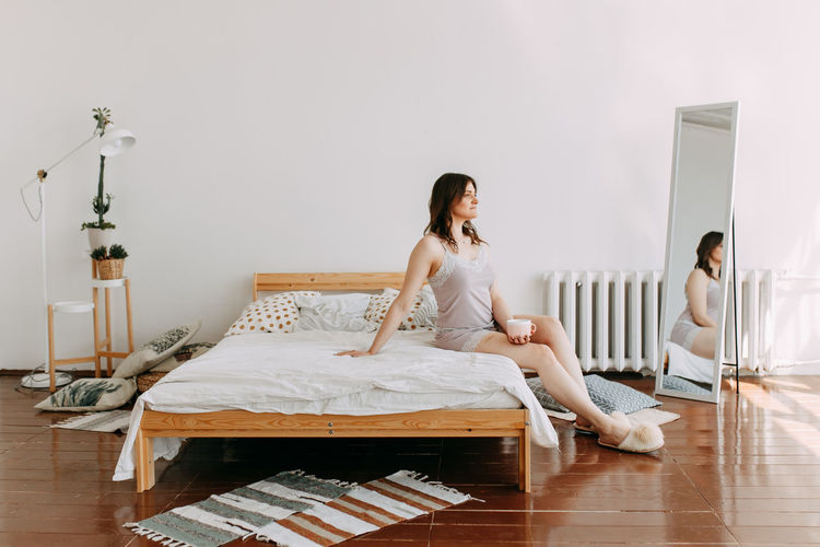 A happy girl in pajamas drinks coffee looks at the phone using technology while lying in bed at home