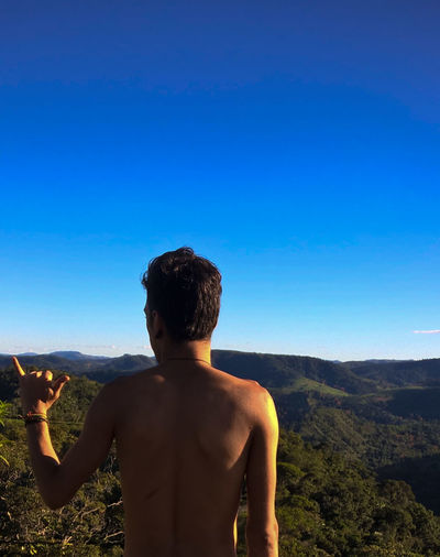 Rear view of shirtless man gesturing shaka sign against clear blue sky