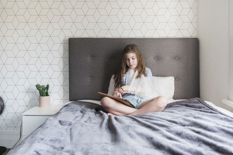 Young girl sitting on bed at home with a broken arm in a sling